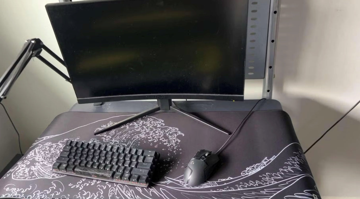 A minimalist computer setup featuring a large monitor, a compact keyboard, and a wired gaming mouse. The items are placed on a large mousepad with a black background and white wave pattern. The monitor is mounted on an adjustable arm, and the overall setup has a clean and organized appearance.