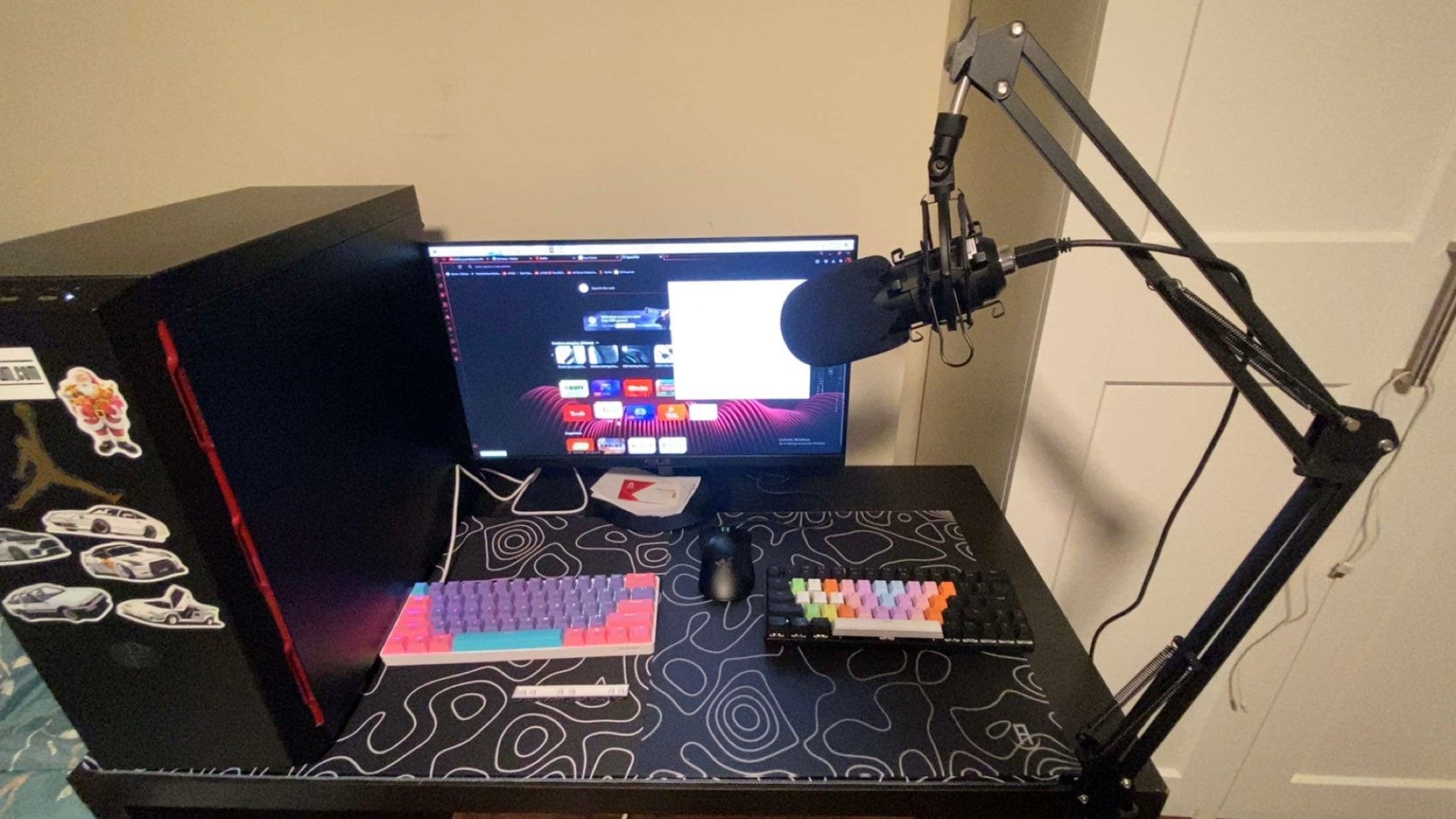 A computer desk setup featuring a monitor displaying a web browser, a black PC tower with various stickers, a microphone on a boom arm, and two mechanical keyboards. The desk is covered with a large mouse pad that has a black topography design with white lines. The keyboard on the left has colorful keycaps, while the one on the right has pastel keycaps.
