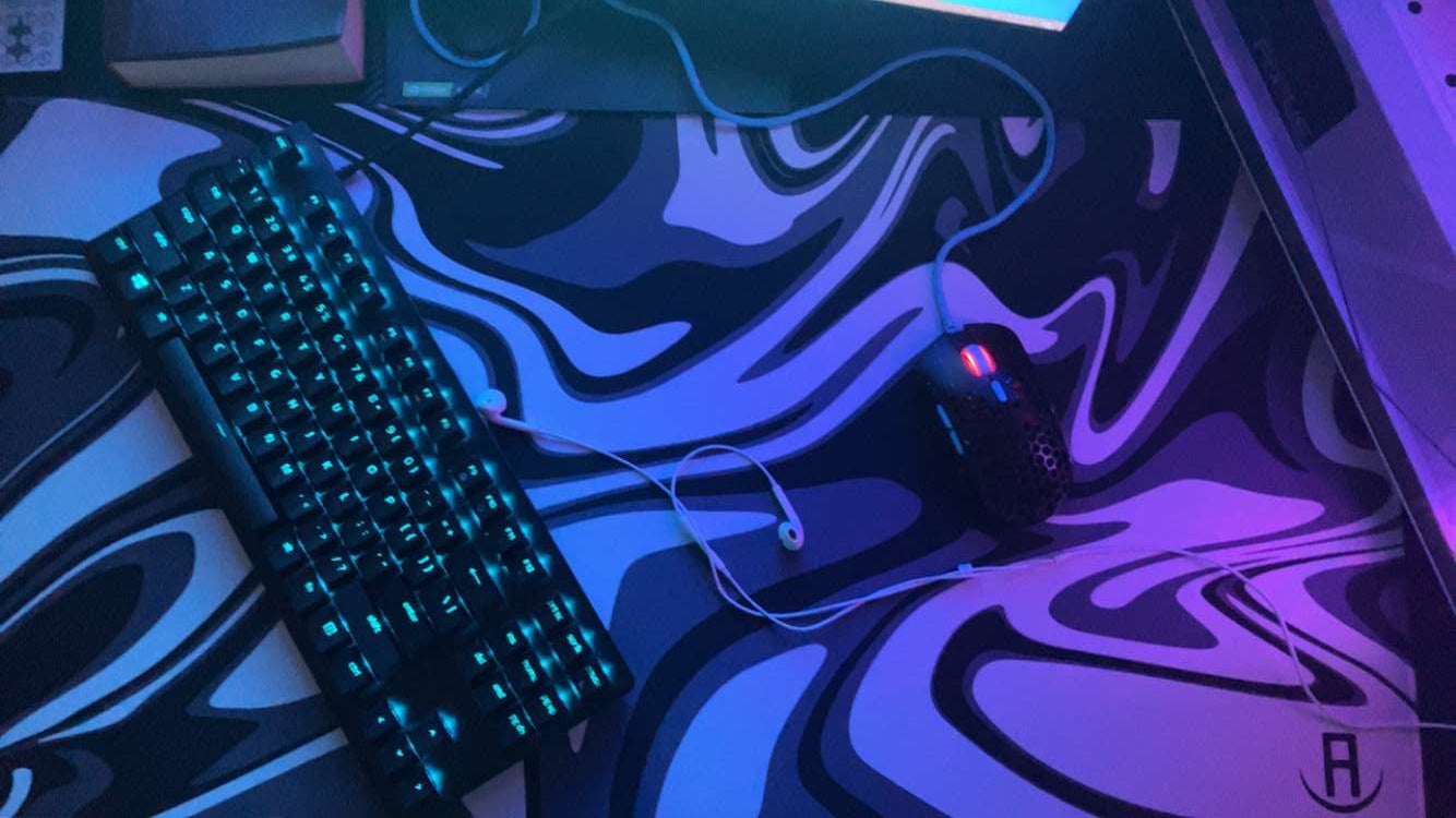 A gaming setup featuring a keyboard with backlit keys emitting a teal glow, a wired mouse with a honeycomb design and red accents, and earphones with a white cable, all placed on a large mousepad with a black and white swirl pattern. The ambient lighting gives the scene a futuristic, neon effect.