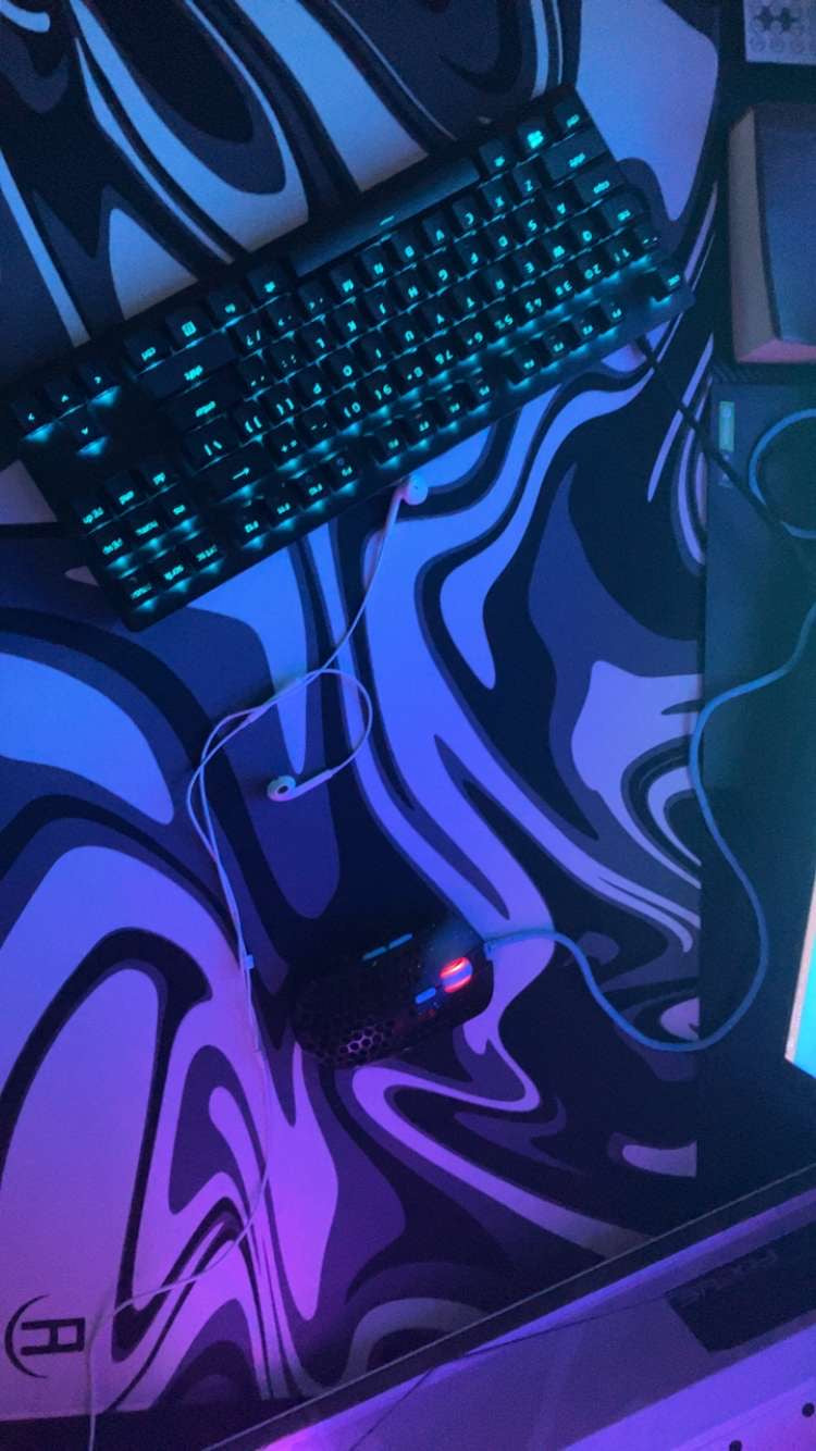 A gaming setup featuring a keyboard with backlit keys emitting a teal glow, a wired mouse with a honeycomb design and red accents, and earphones with a white cable, all placed on a large mousepad with a black and white swirl pattern. The ambient lighting gives the scene a futuristic, neon effect.