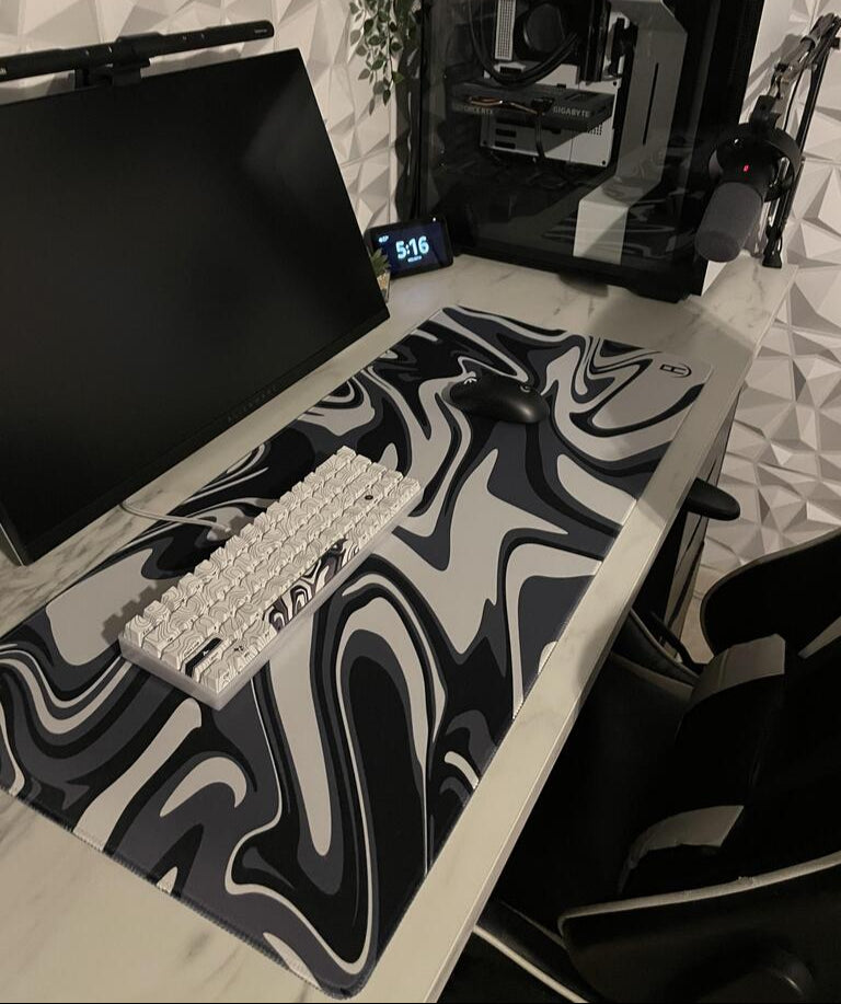 A sleek gaming setup featuring a black monitor, a black and white mechanical keyboard, and a black wireless mouse on a large gaming mouse pad with a black, white, and grey marble pattern. 