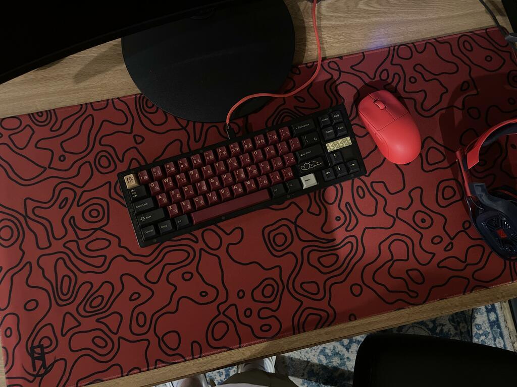 A desk setup featuring a red and black topography-patterned mousepad that covers a large portion of the desk. On top of the mousepad, there is a mechanical keyboard with black and red keycaps, a red mouse, and a pair of headphones. The mousepad has an abstract, wavy topographic design with black lines on a red background.